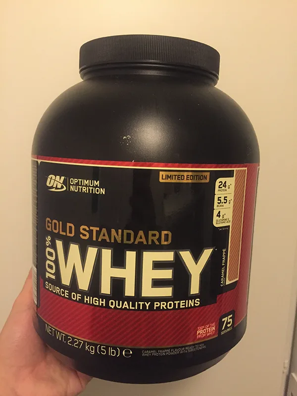 Product photo of a caramel frappe protein powder tub beside a branded serving glass.